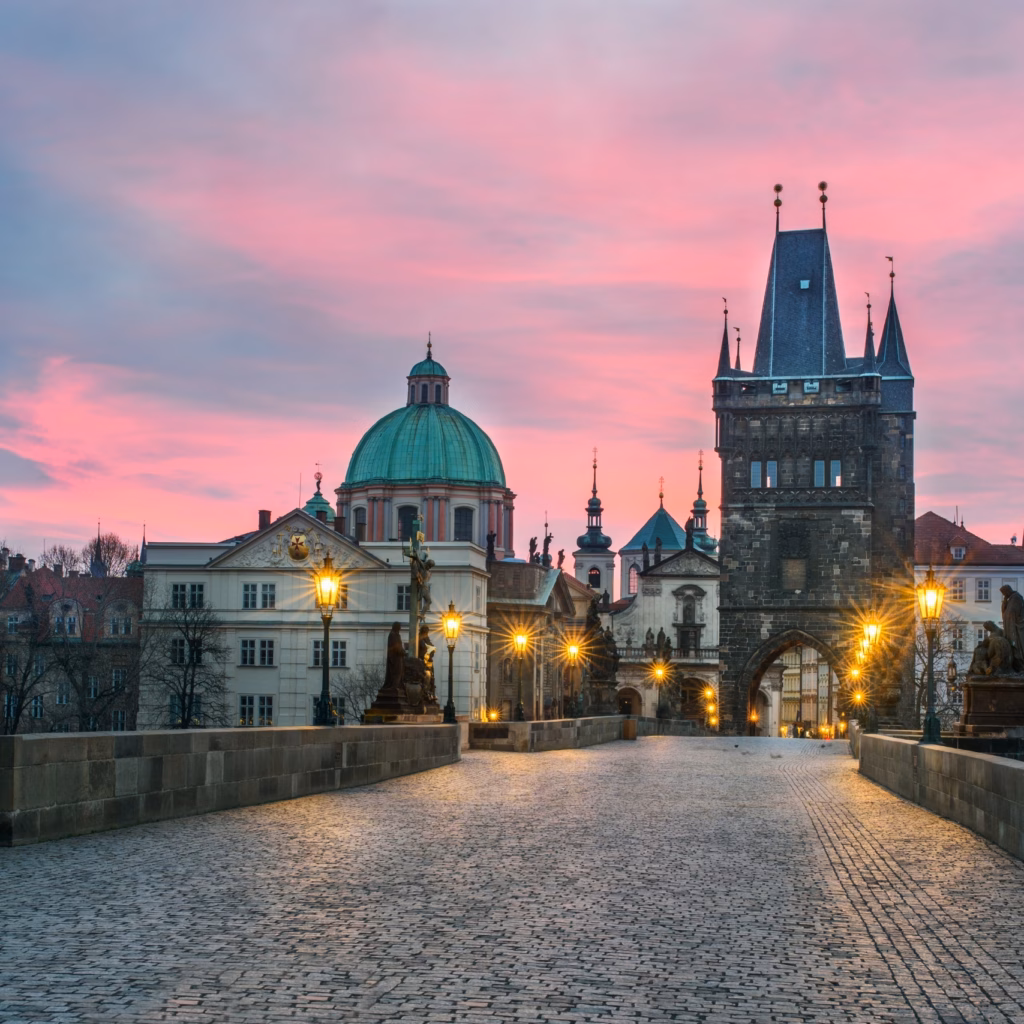 The historic Charles Bridge in Prague lit by warm street lamps at dusk, with Baroque statues and Gothic towers under a pink and blue sky.